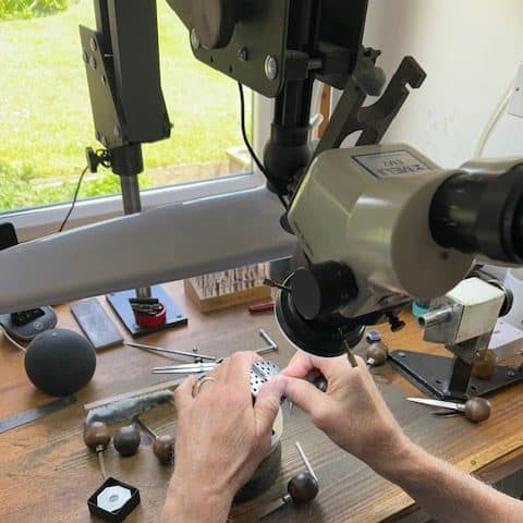 A wide shot of a traditional jeweler's bench showing a specialist hand engraver at work under a microscope, surrounded by wooden-handled steel engraving tools, dividers, and a wax impression mold.