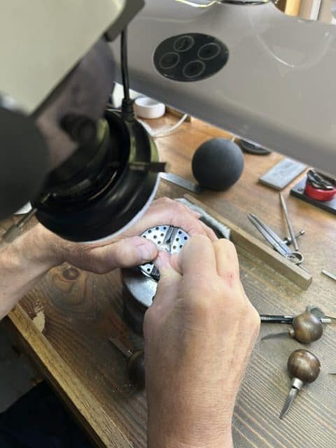 An overhead view of a master engraver working under a professional bench microscope, carefully sculpting a heraldic design on a custom gold ring at a wooden jeweler's bench.