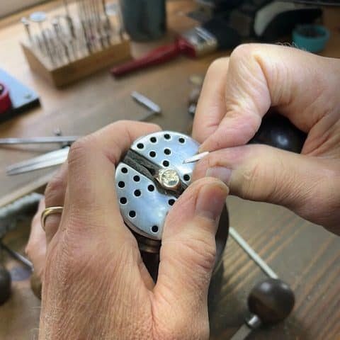 Close-up of a specialist seal hand engraver using a steel graver tool to precisely carve an intricate design into a gold signet ring held securely in an engraver's ball vise.
